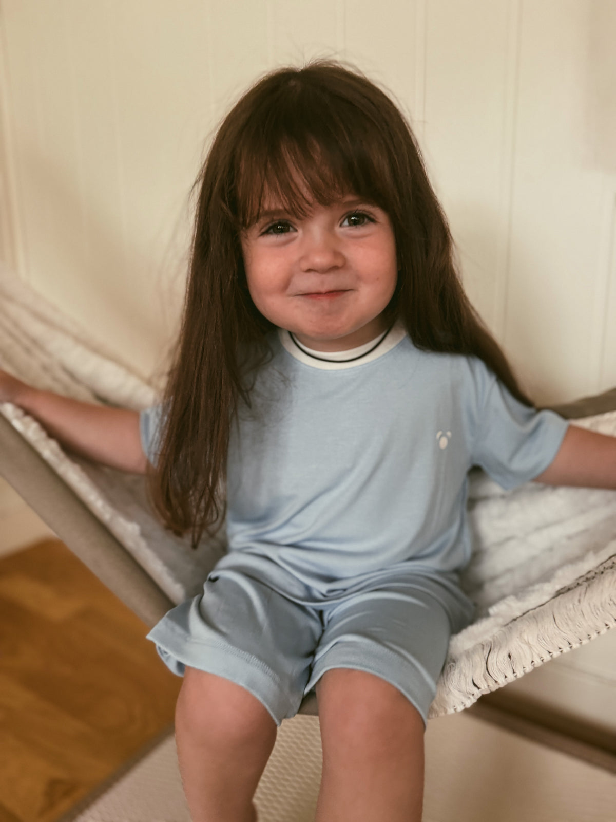 A young child with long brown hair sits in a fabric hammock wearing a light blue short-sleeve t-shirt and matching shorts. She is wearing Fluffy Panda bamboo pyjamas in Cornflower. The background shows a cream-colored paneled wall and a wooden floor.